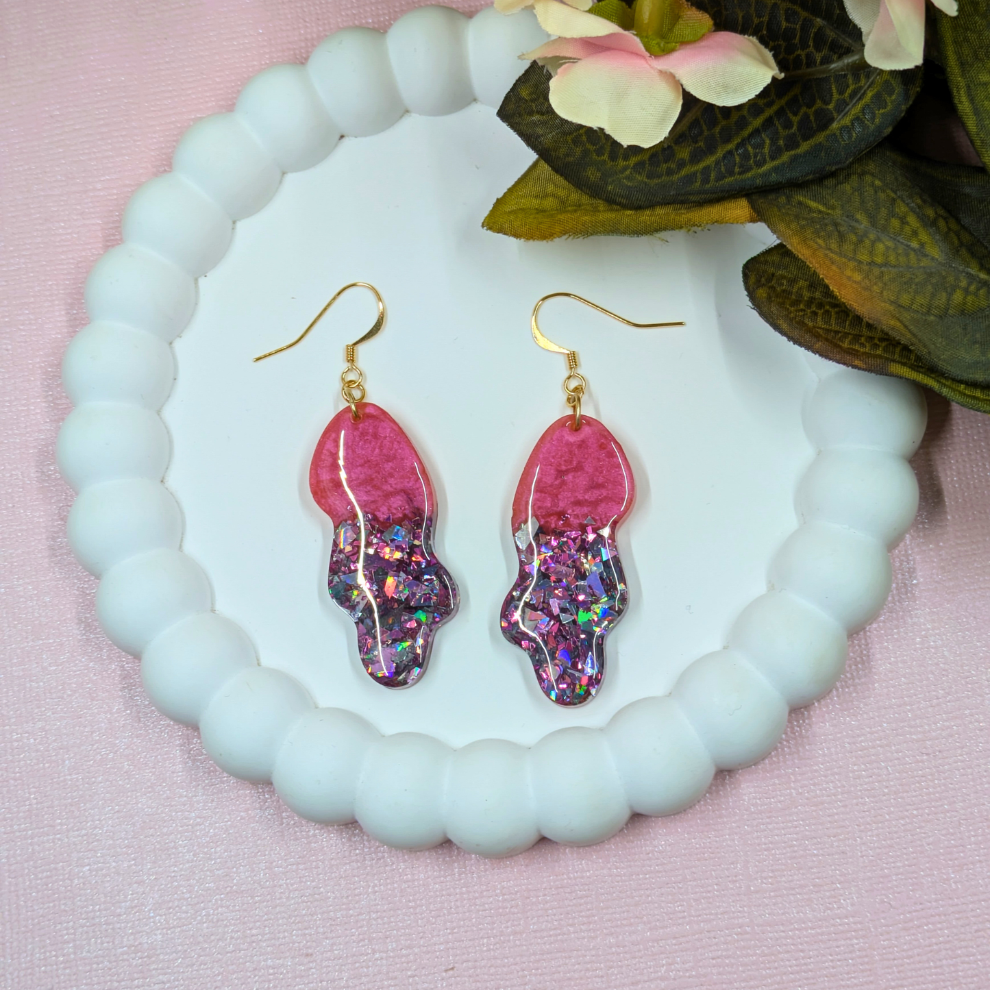 Pink glittery earring with gold hooks on a light blue stand with flowers in the background