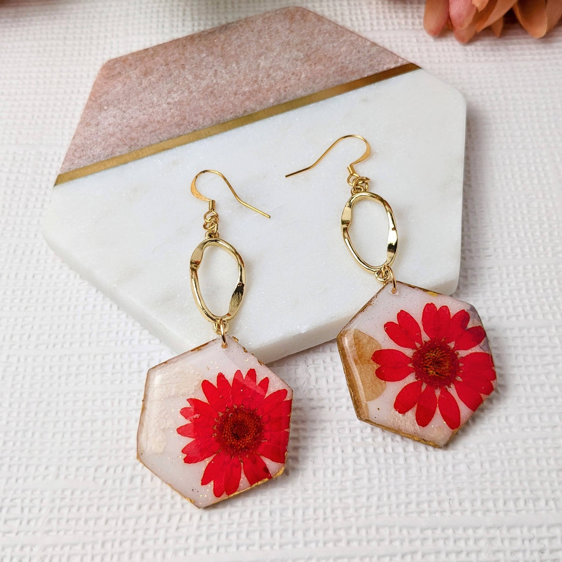 A pair of red flower dangle earrings with gold-colored hexagon shapes and gold leaf detailing, on a white surface with a pink flower in the background.