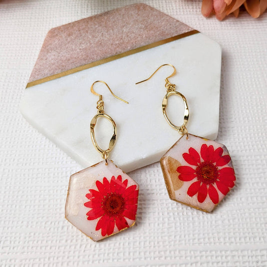 A pair of red flower dangle earrings with gold-colored hexagon shapes and gold leaf detailing, on a white surface with a pink flower in the background.