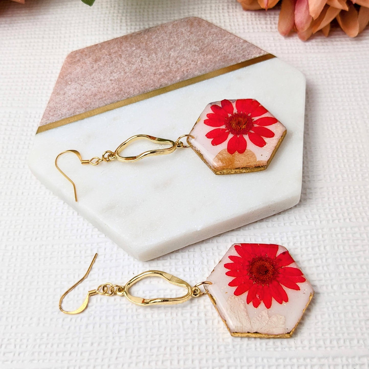Hexagonal earrings with red floral designs on a marble surface with flowers in the background