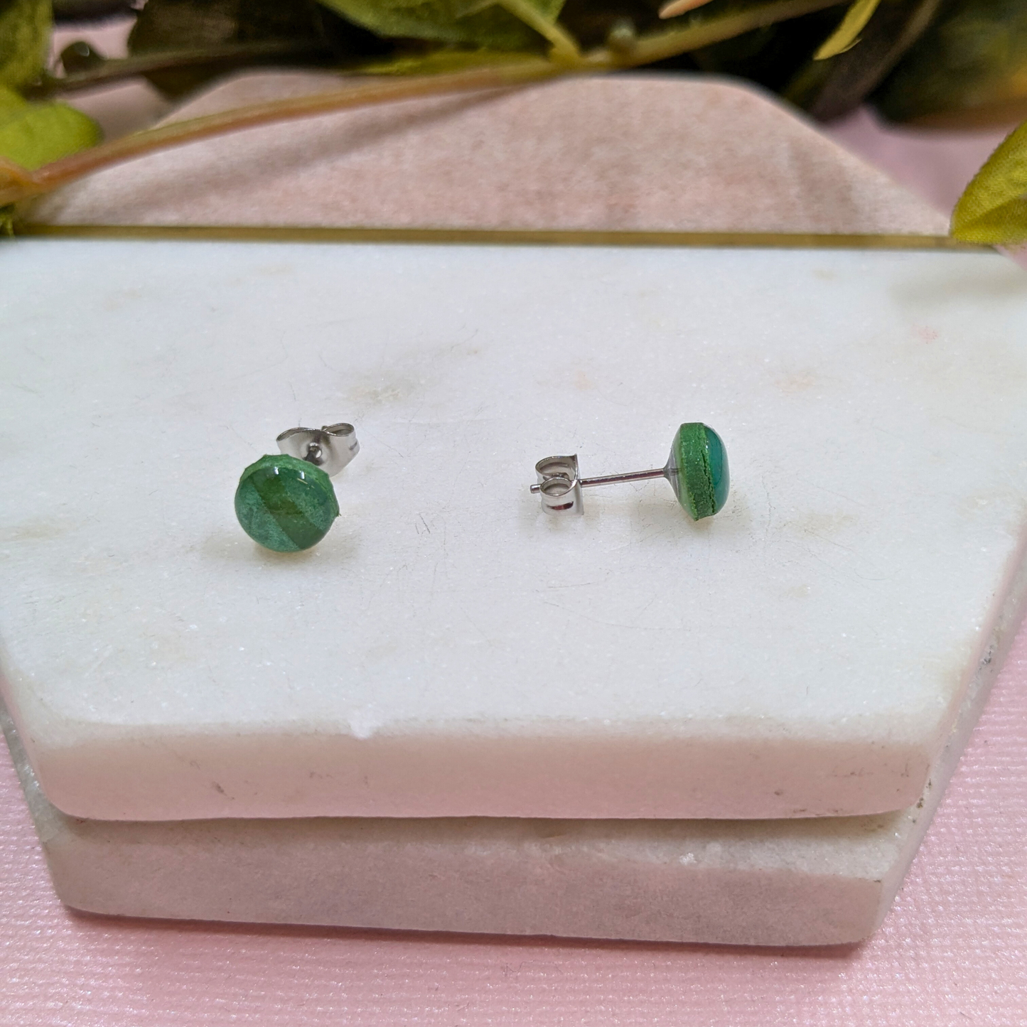 Green stud earrings on a white marble surface with green leaves in the background