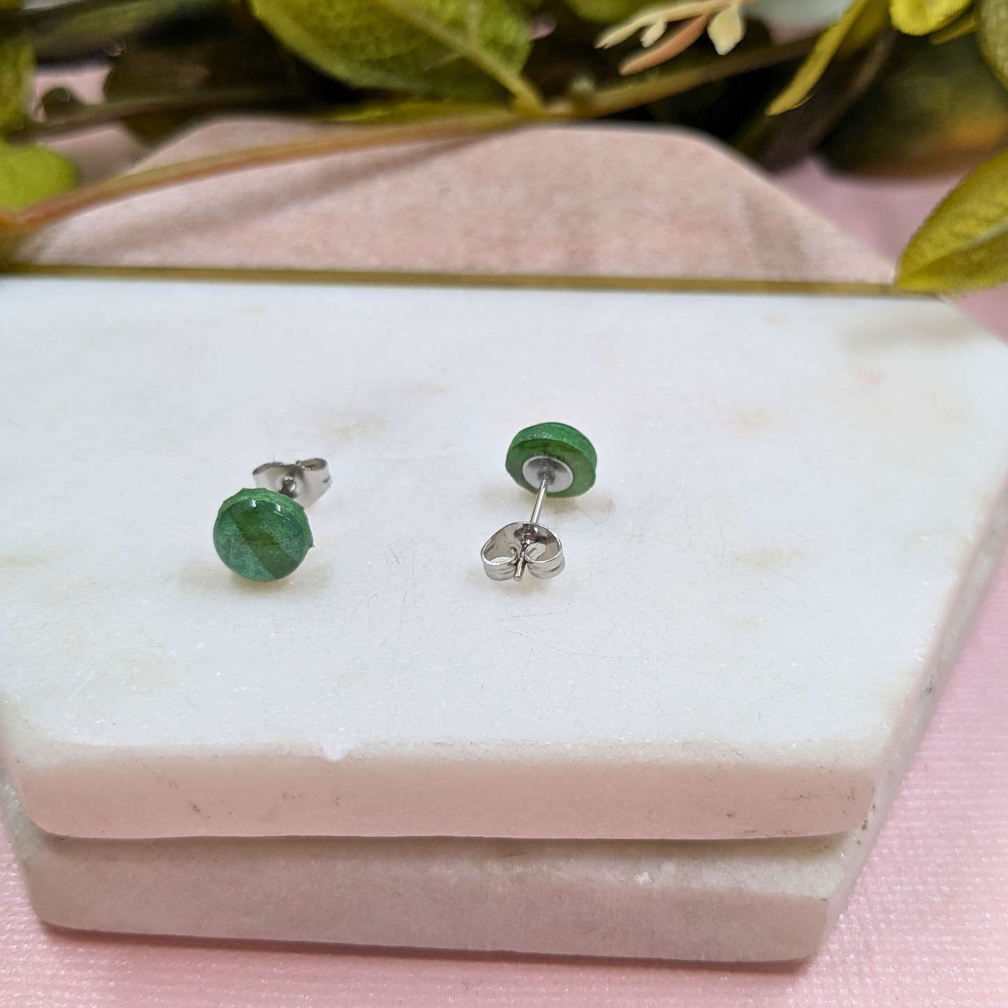 Green stud earrings on a white jewelry box with pink surface and green leaves in the background