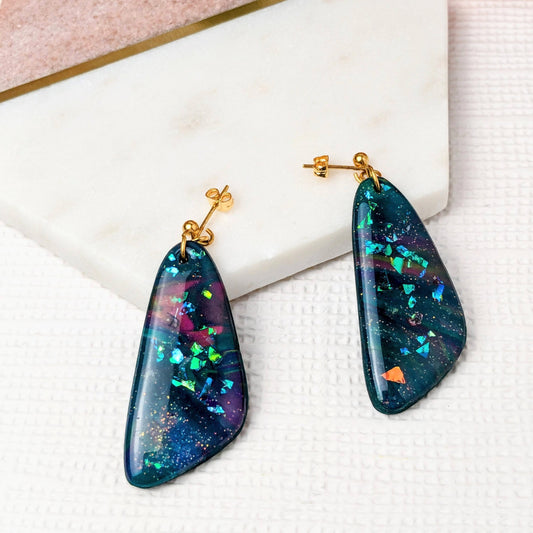 A pair of blue iridescent polymer clay dangle earrings with gold-plated ball stud ear posts, displayed on a white surface with floral accents.