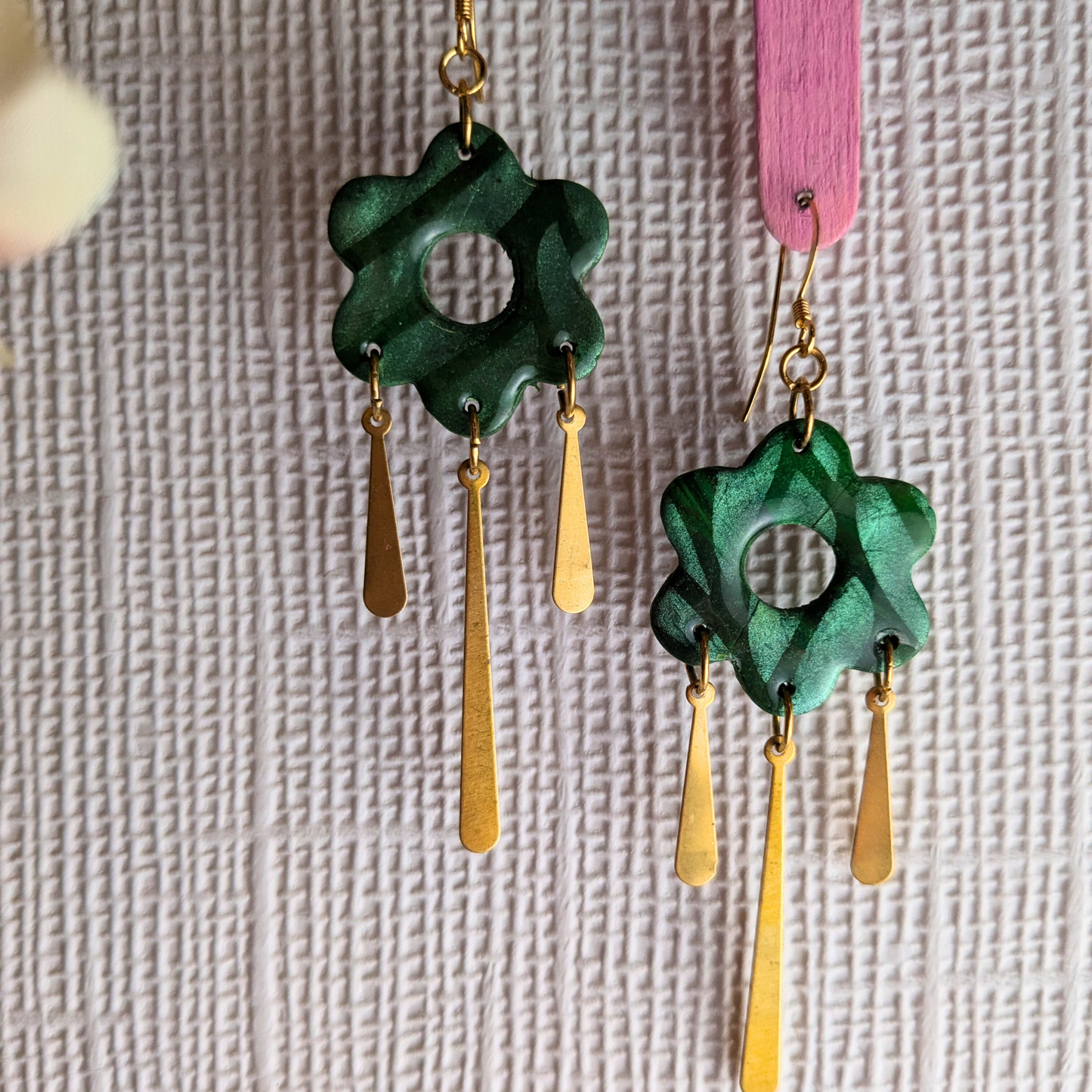 A pair of handmade dangle earrings featuring textured dark green flower-shaped pendants with gold-tone hooks and three dangling gold-tone teardrop charms, shown against a textured light gray background.