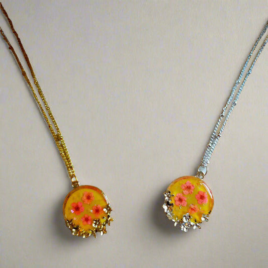 A gold-plated necklace featuring a resin flower pendant with pink blossoms and a yellow background, displayed on a textured surface with a white flower in the background.