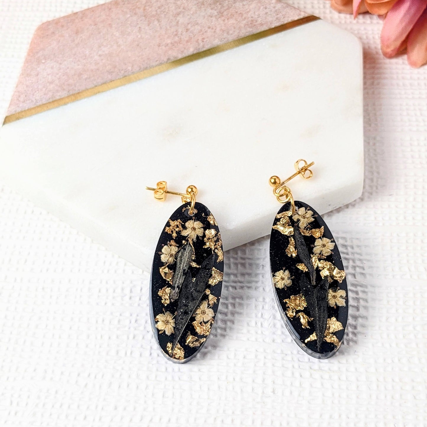 A pair of oval dangle earrings featuring black resin with gold leaf and floral patterns, displayed on a white surface with pink flowers in the background.
