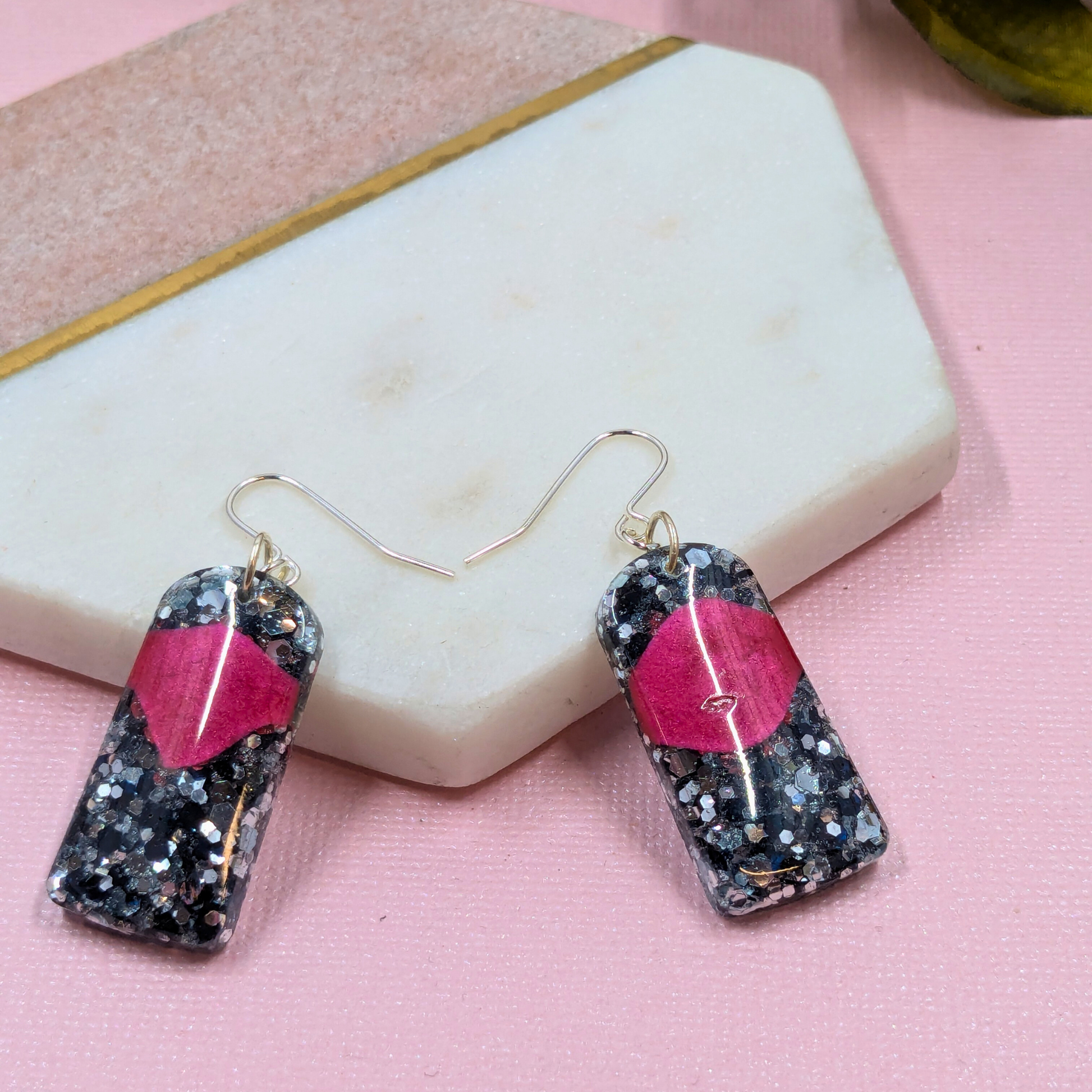 Earrings with black and silver glitter and a red accent rest on a hexagonal stone backdrop. The scene has a stylish, modern tone.