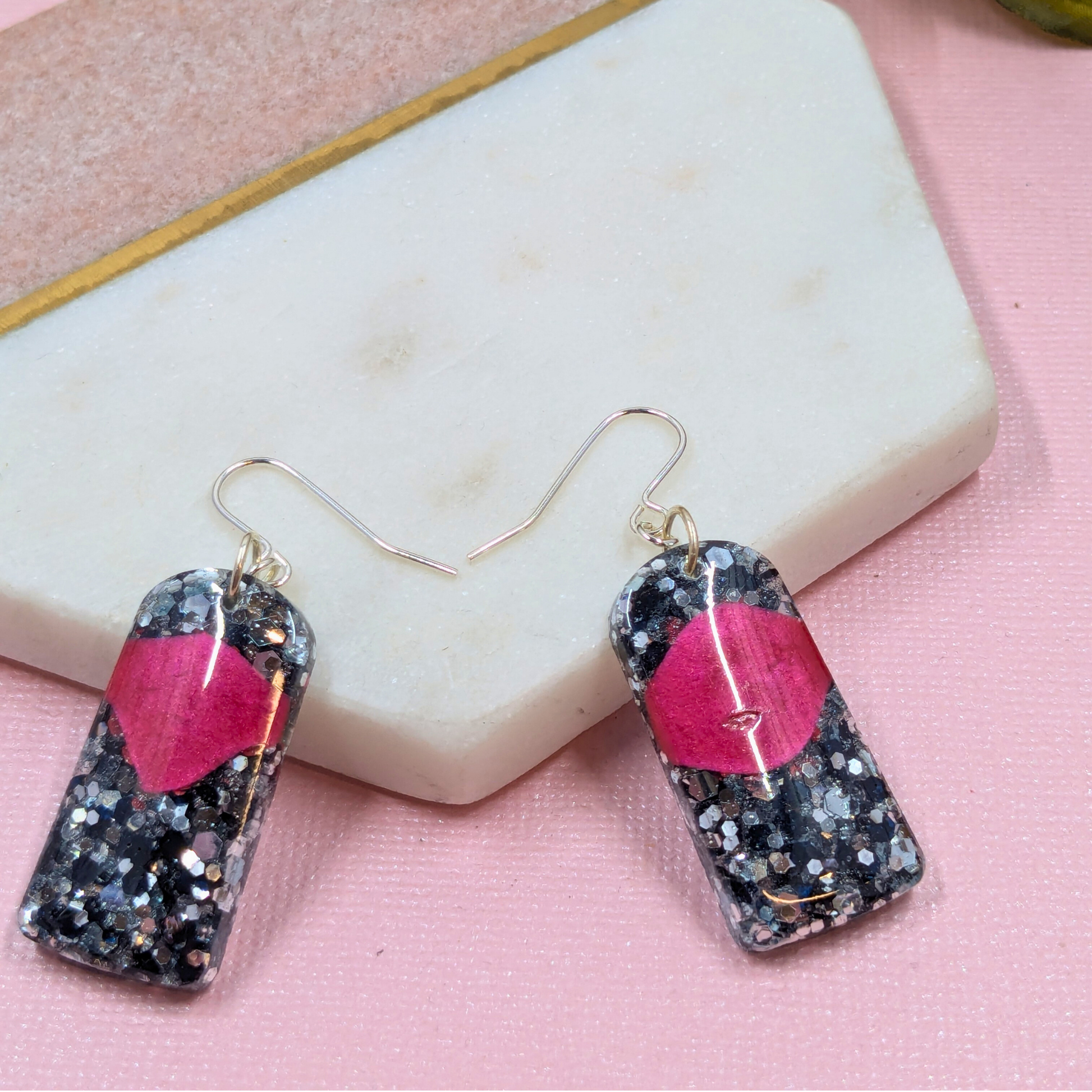 Earrings with black and silver glitter and a red accent rest on a hexagonal stone backdrop. The scene has a stylish, modern tone.