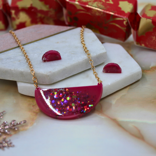 A gold chain necklace with a semi-circular magenta pink pendant filled with iridescent glitter, displayed on stacked white stones with matching pink earrings and red gift boxes in the background.