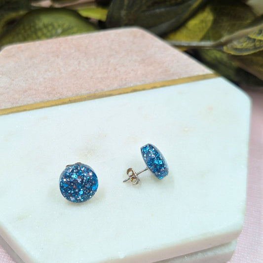 Blue glittery stud earrings on a white marble surface with green leaves in the background