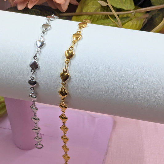 Two heart-link bracelets, one silver-toned and one gold-toned, displayed on a white cylindrical jewelry stand.