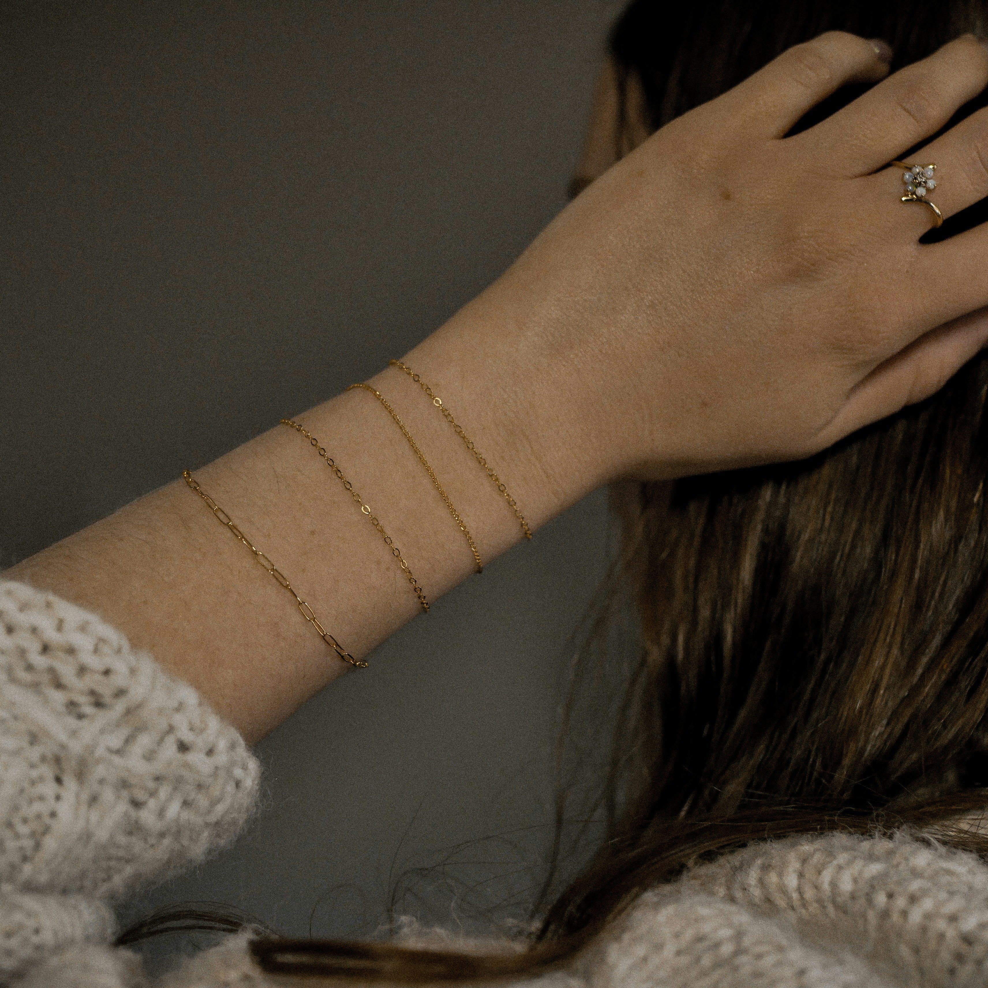 A person with light skin touches their hair, wearing a permanent gold bracelet and a ring with a small, clear stone. They are dressed in a textured sweater.