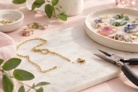 A broken gold chain and loose clasp on a marble jewellery workbench, beside pliers and a dish of colourful resin pieces being reworked into new jewellery