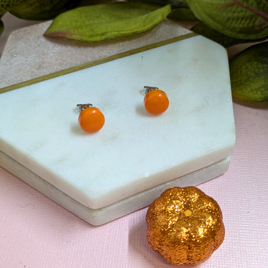 A pair of small orange round stud earrings with silver posts, displayed on a white marble hexagonal tray next to a glittery gold pumpkin prop on a pink surface.