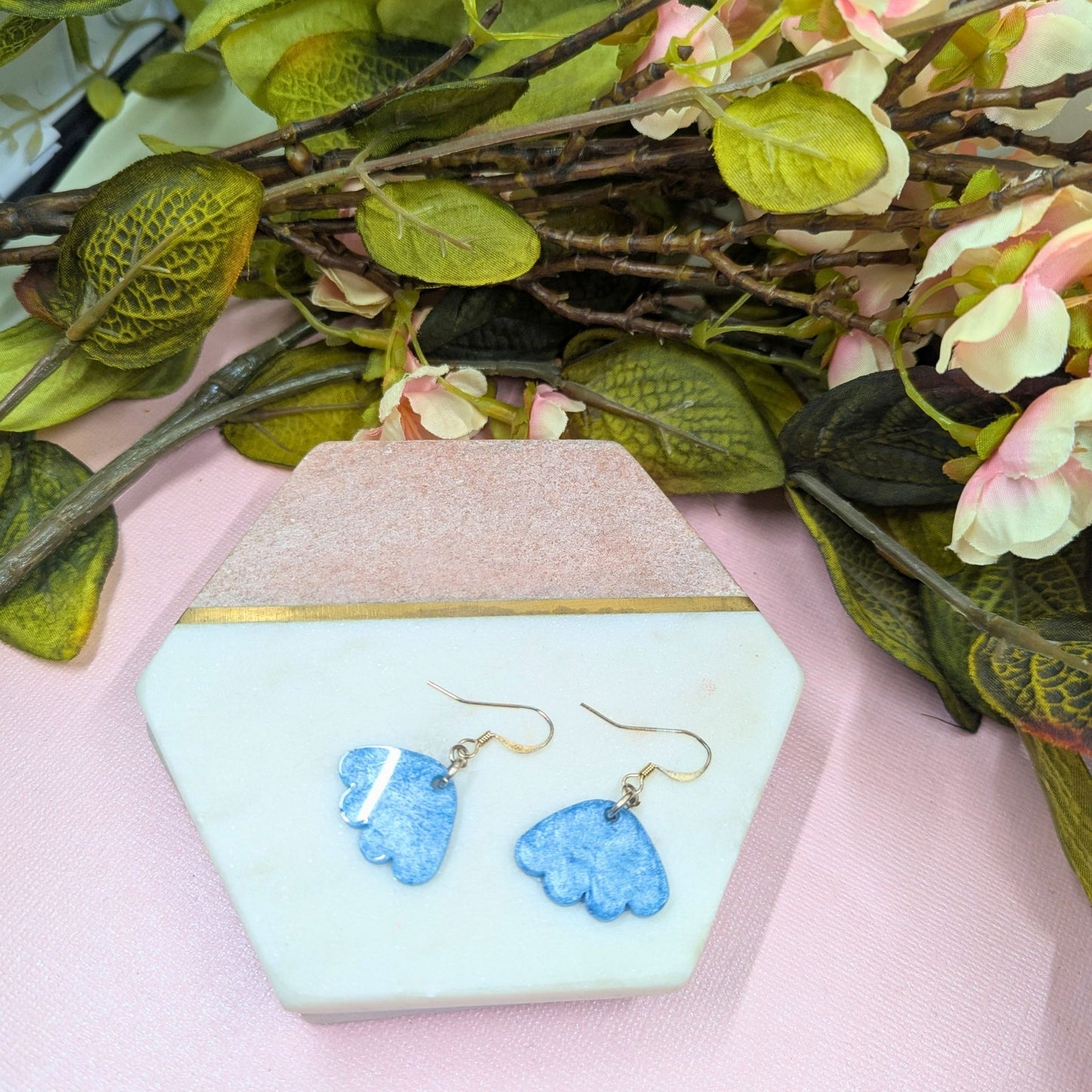 Blue floral earrings on a hexagonal green and gold jewelry holder with pink background and green leaves.