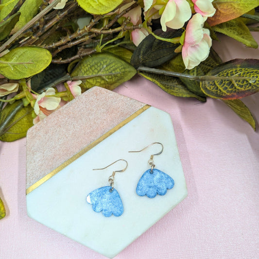 Blue flower-shaped earrings on a decorative stand with greenery and flowers in the background