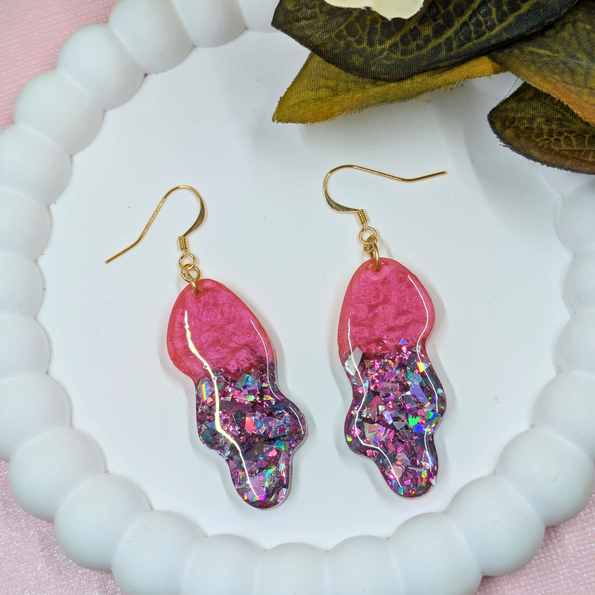Pink glittery earrings on a white and pink textured surface with leaves.