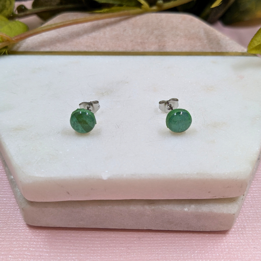 Green stud earrings on a white marble surface with a pink fabric background