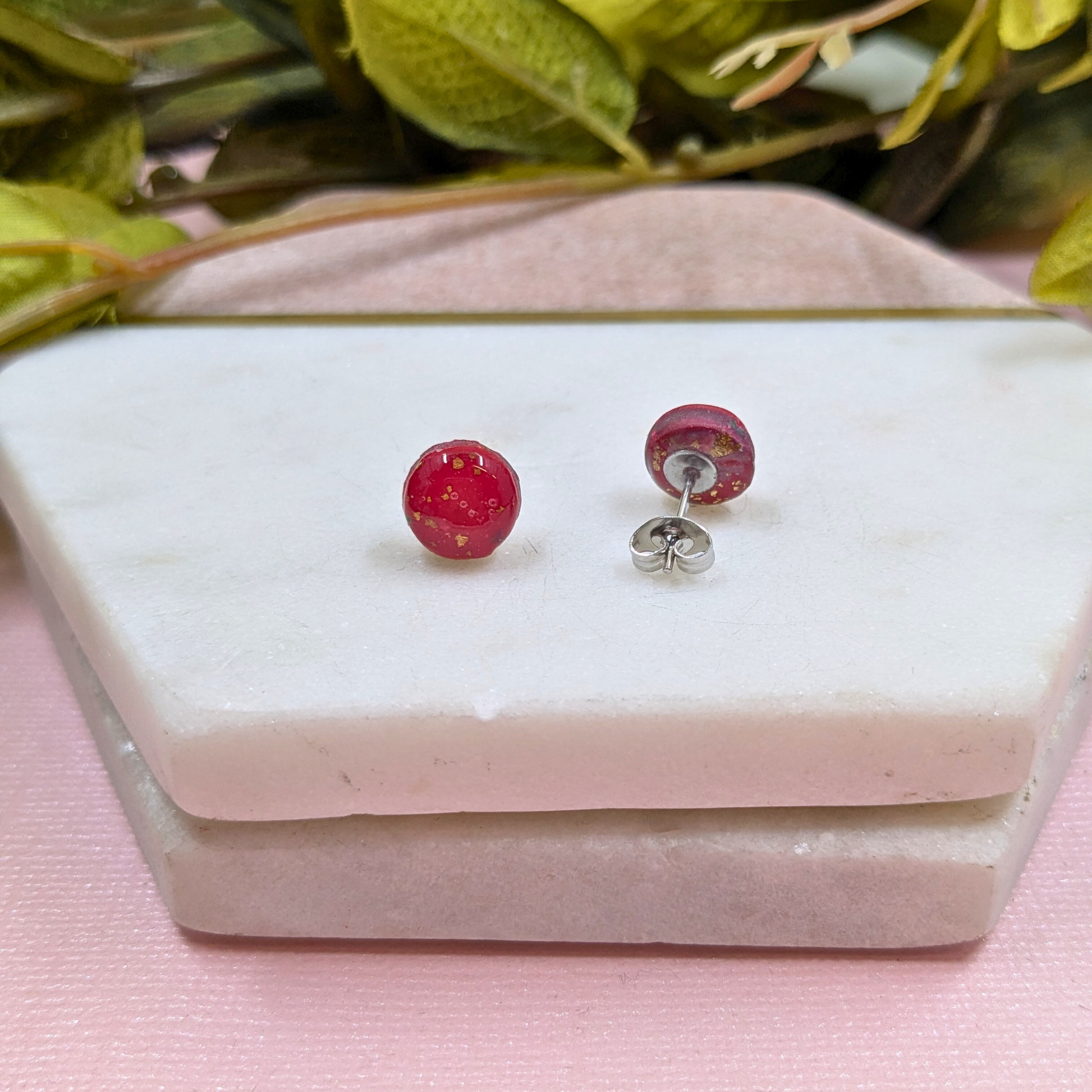 Red berry stud earrings on a white stone surface with green leaves in the background