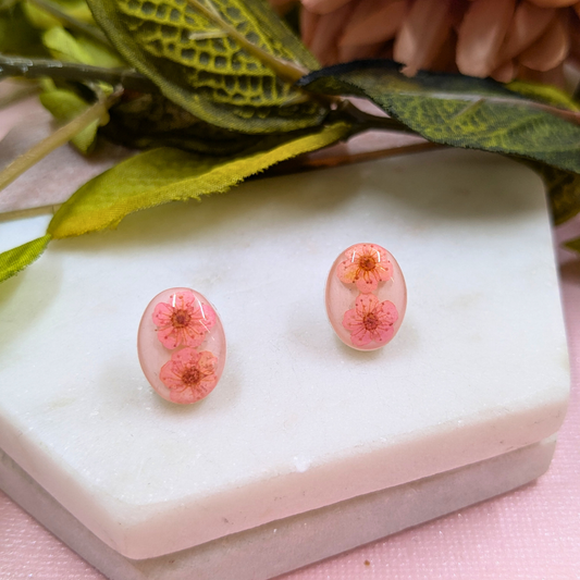 A pair of oval stud earrings with pink cherry blossom flowers encased in clear resin, displayed on a white stone block with green leaves in the background.