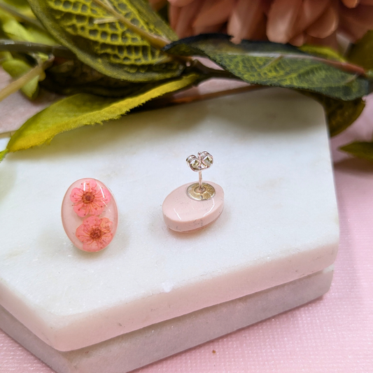 A pair of small oval stud earrings displayed on a pale pink and white block. One earring is front-facing, showing a translucent pink resin face with two pressed pink flowers; the other is turned sideways, showing a silver-toned post and backing.
