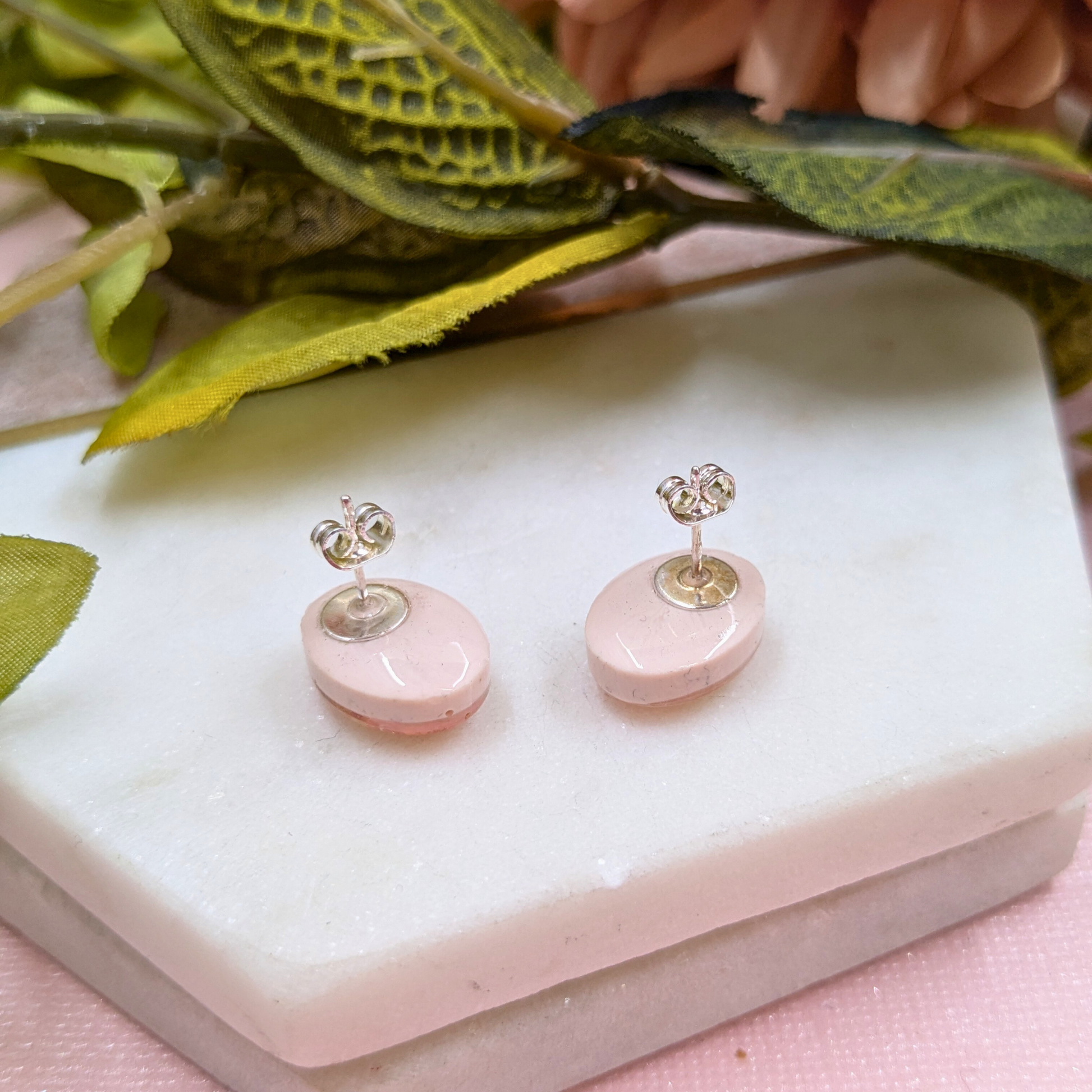 A pair of small stud earrings with oval pale pink stone discs and silver posts, displayed on a white rectangular block with decorative green leaves and pink flowers in the background.