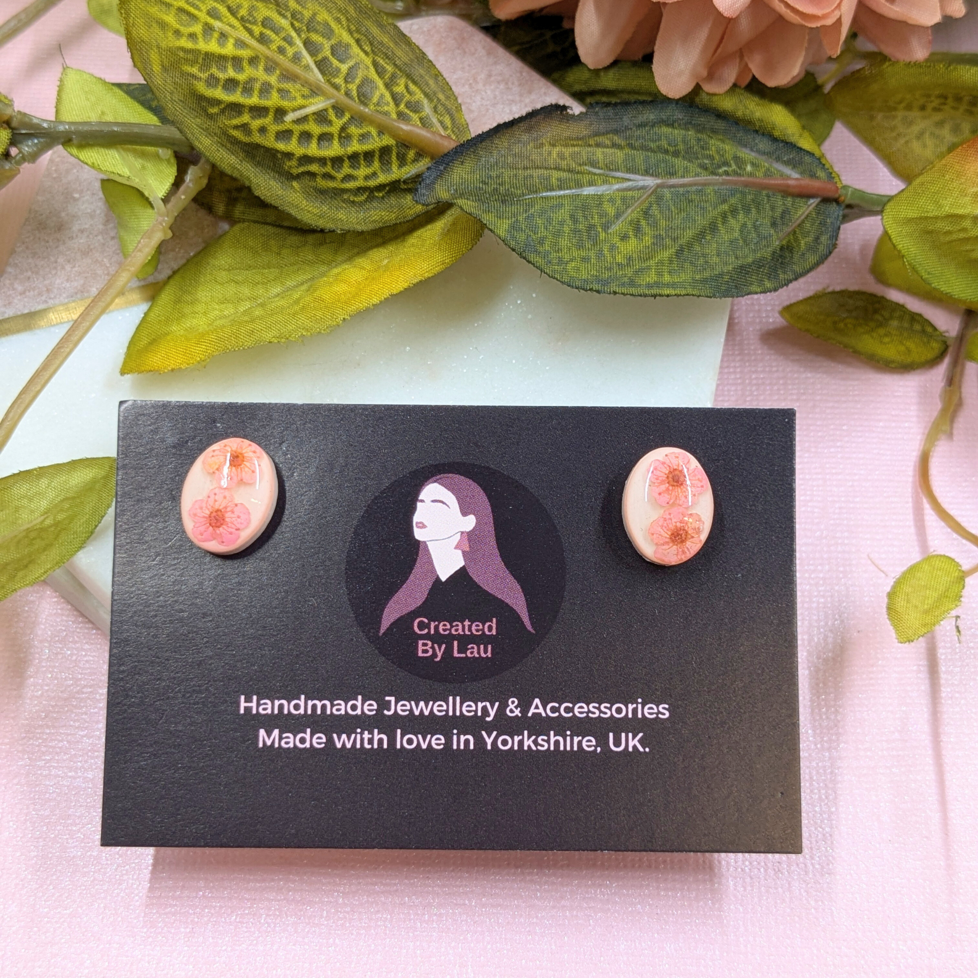 A pair of handmade oval stud earrings with pink and peach floral designs, displayed on a black card labeled 'Created By Lau' with the text 'Handmade Jewellery & Accessories Made with love in Yorkshire, UK.' The card is styled with green leaves and pale pink flowers on a light pink surface.