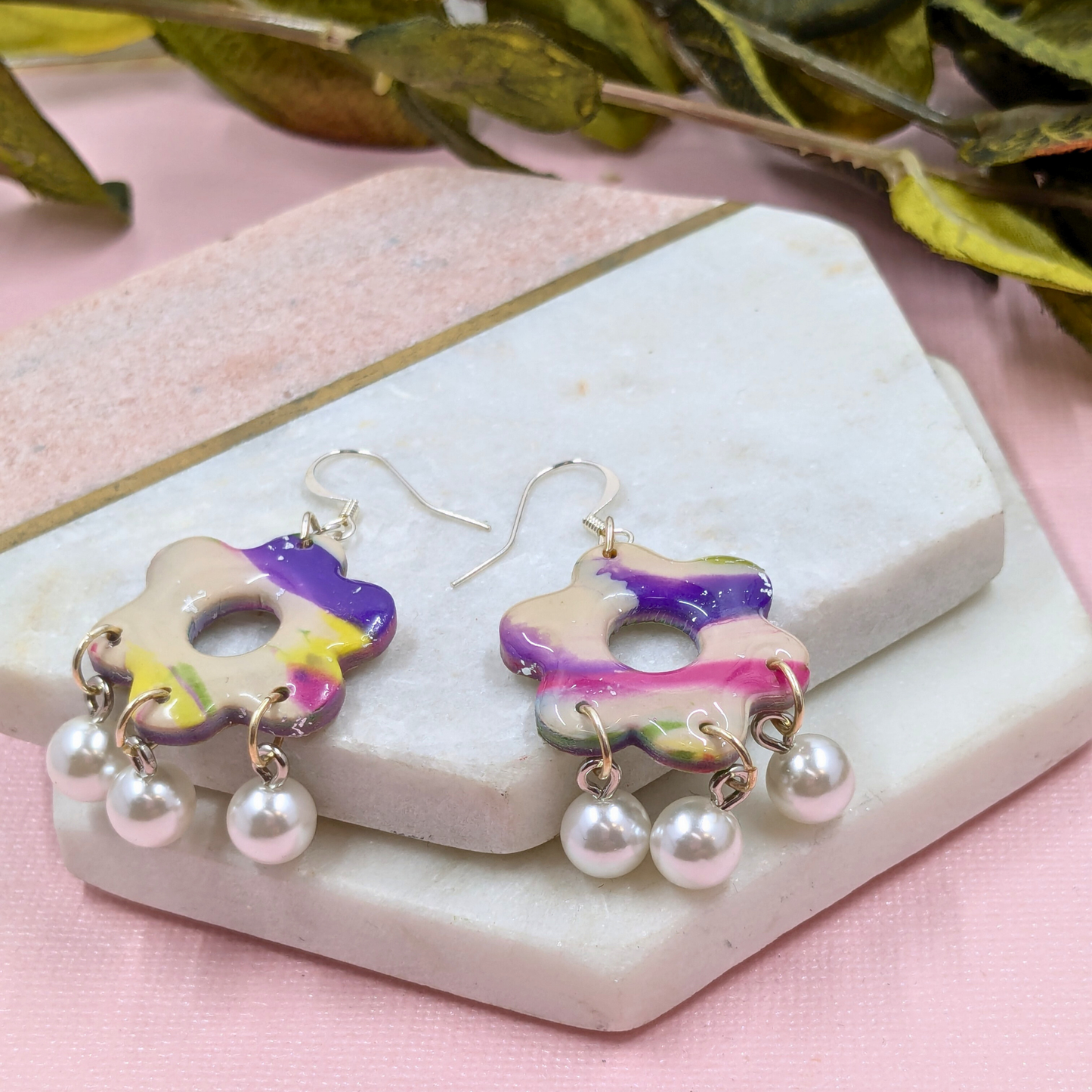 Colourful flower-shaped polymer clay earrings with marbled purple, pink, and yellow hues, adorned with dangling pearls, on a textured stone background.