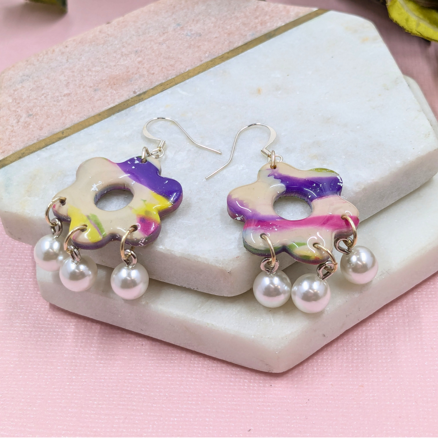 Colourful flower-shaped polymer clay earrings with marbled purple, pink, and yellow hues, adorned with dangling pearls, on a textured stone background.