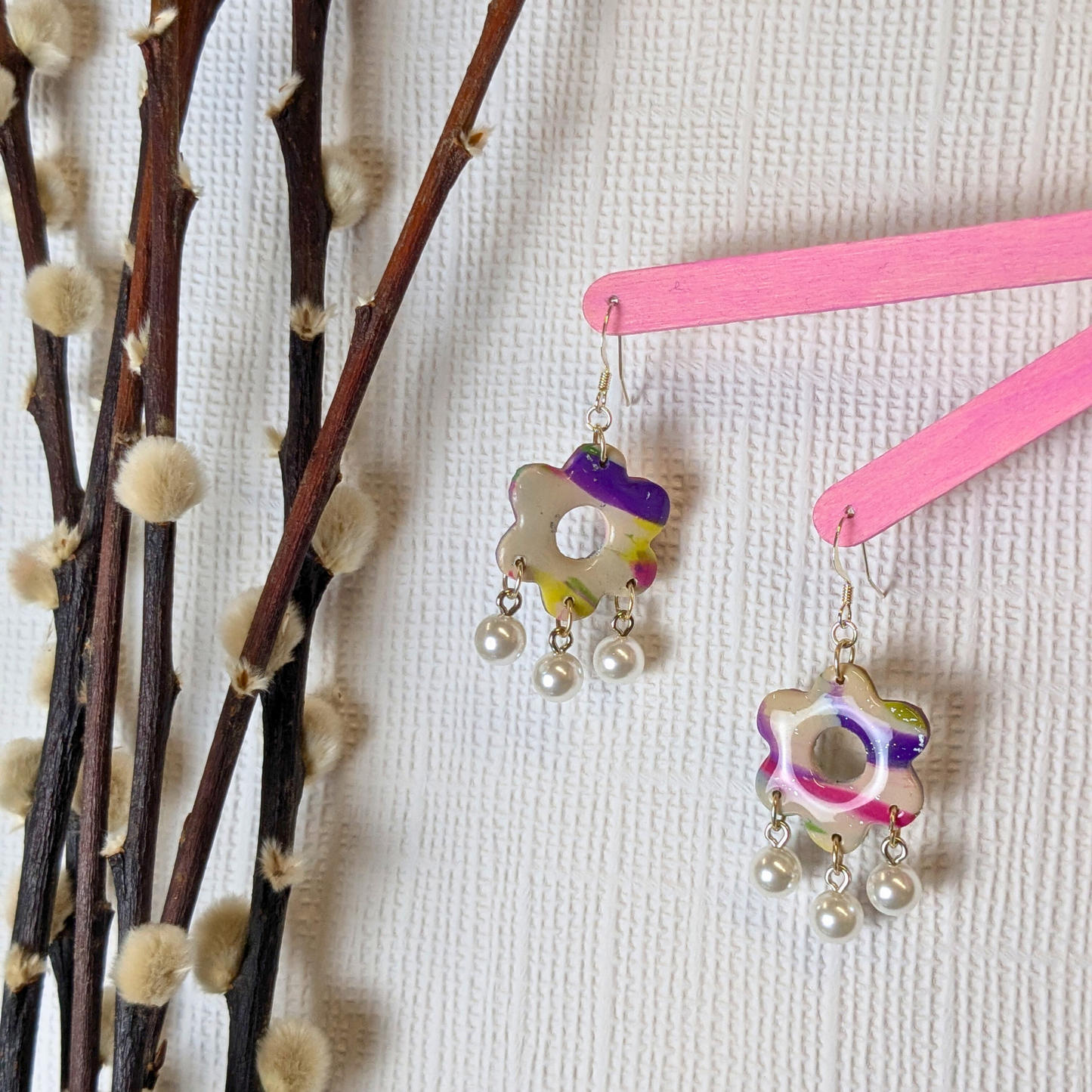 Colourful floral polymer clay earrings with pearl drops hang on pink sticks against a textured white background, beside fluffy branches, creating a whimsical vibe.