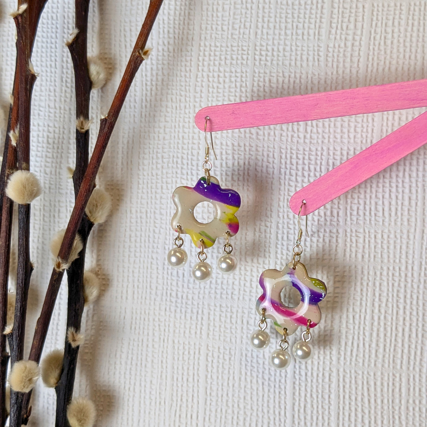 Colourful floral polymer clay earrings with pearl drops hang on pink sticks against a textured white background, beside fluffy branches, creating a whimsical vibe.