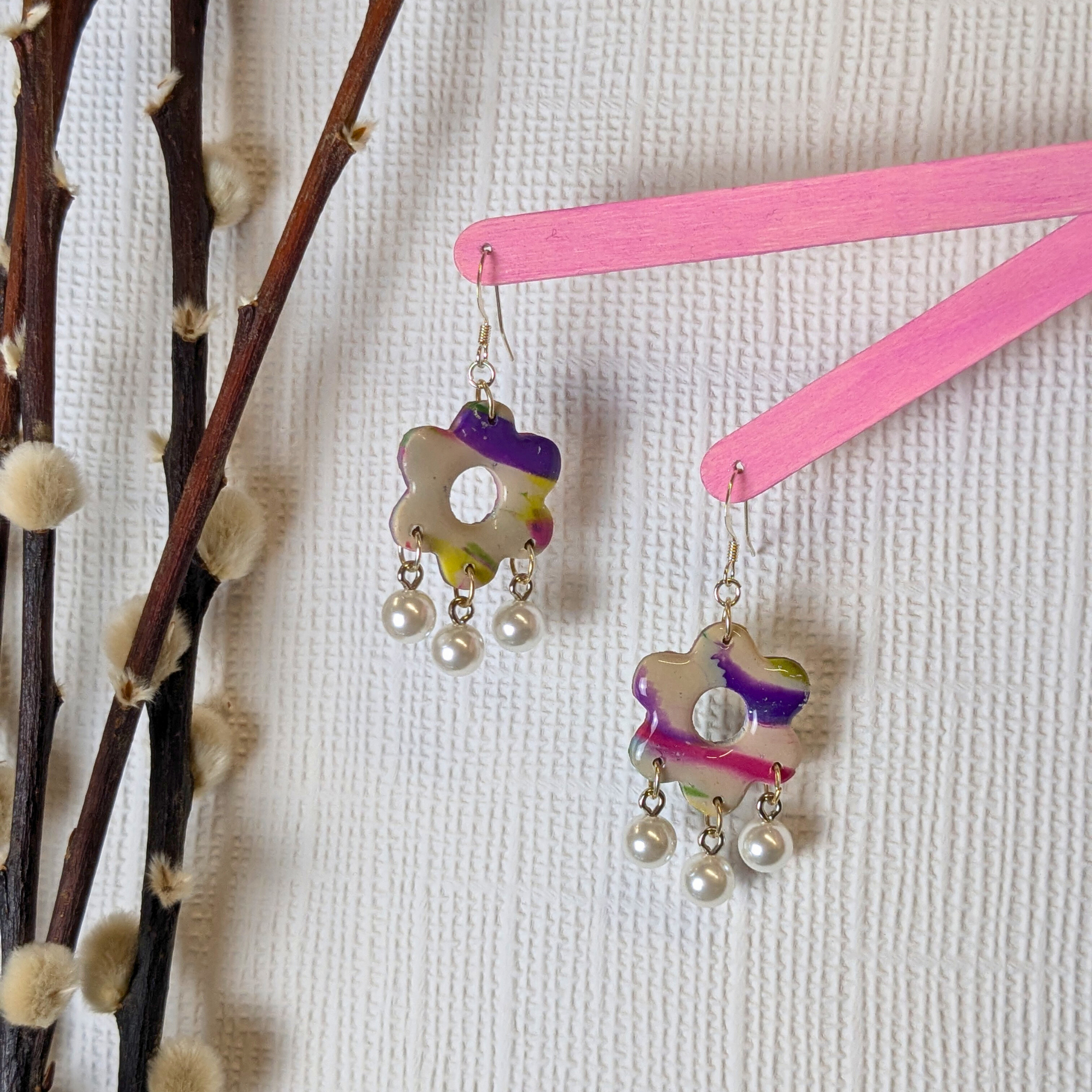 Colourful floral polymer clay earrings with pearl drops hang on pink sticks against a textured white background, beside fluffy branches, creating a whimsical vibe.