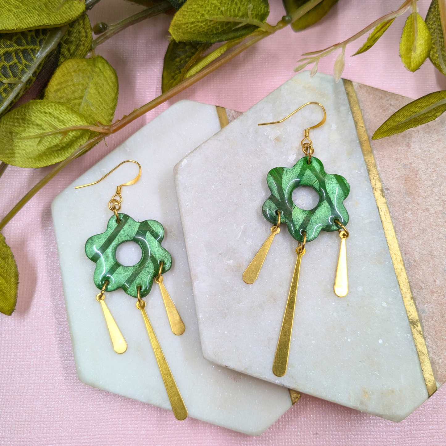A pair of dangling earrings displayed on white stone slabs over a pink fabric background with green leaves. The earrings feature a green marbled flower-shaped charm with a gold fishhook and two gold teardrop drops.