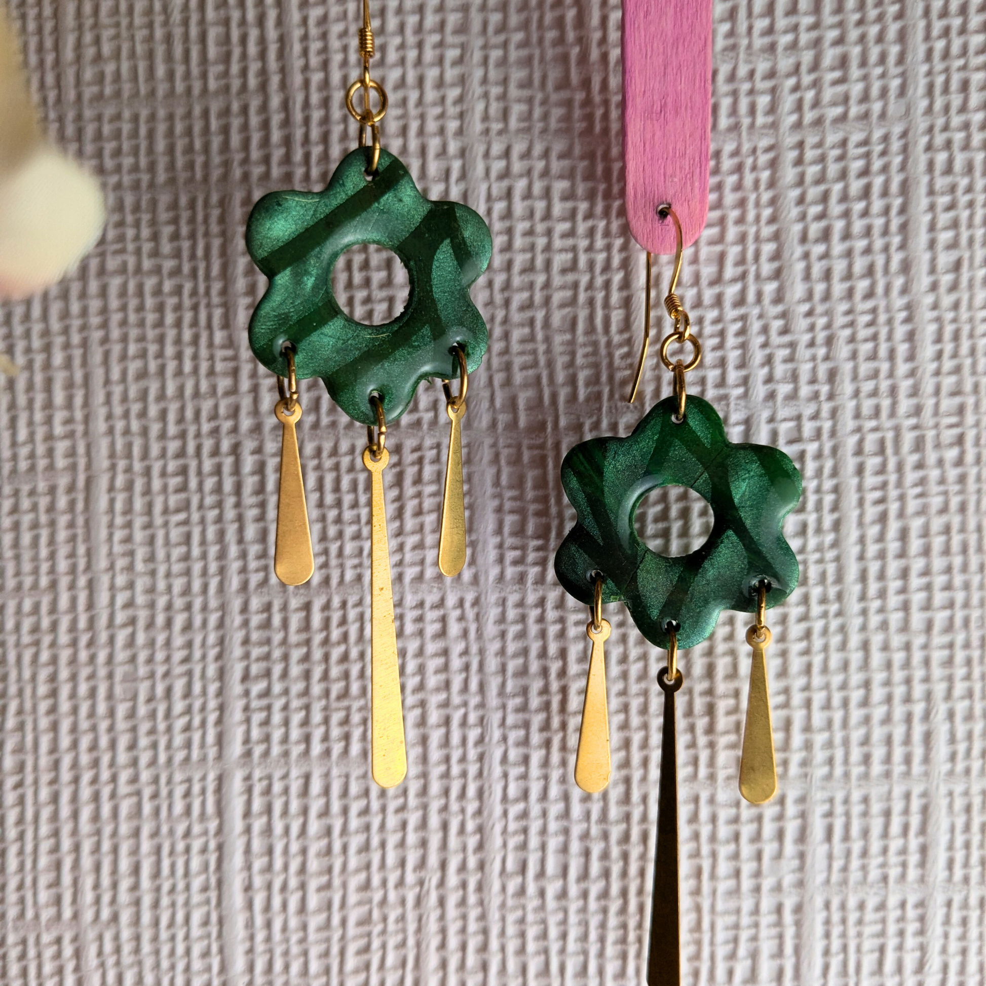 A pair of handmade dangle earrings featuring green flower-shaped charms with gold-toned metal wire details and hanging gold-toned teardrop charms, shown against a textured light background.