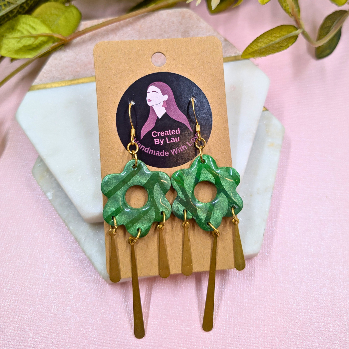 A pair of handmade dangle earrings displayed on a kraft paper card. The earrings feature textured green flower-shaped charms with gold-tone hooks and dangling gold-tone bar charms.