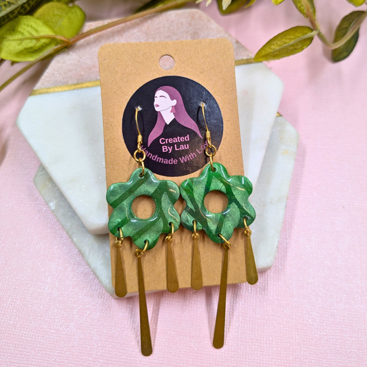 A pair of handmade dangle earrings displayed on a kraft paper card. The earrings feature textured green flower-shaped charms with gold-tone hooks and dangling gold-tone bar charms.