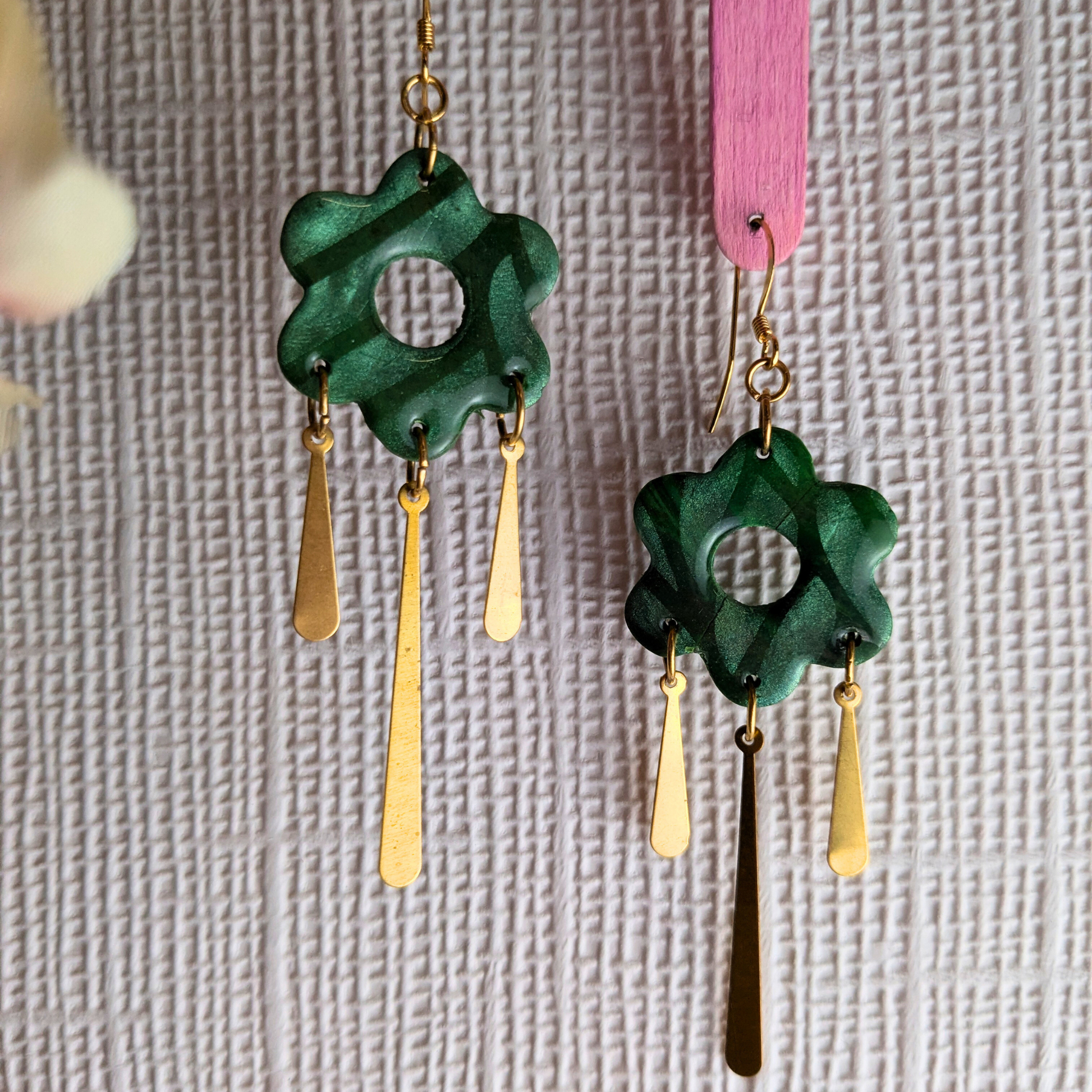 A pair of handmade dangle earrings with dark green flower-shaped charms and gold-tone teardrop charms, hanging against a textured light gray background.