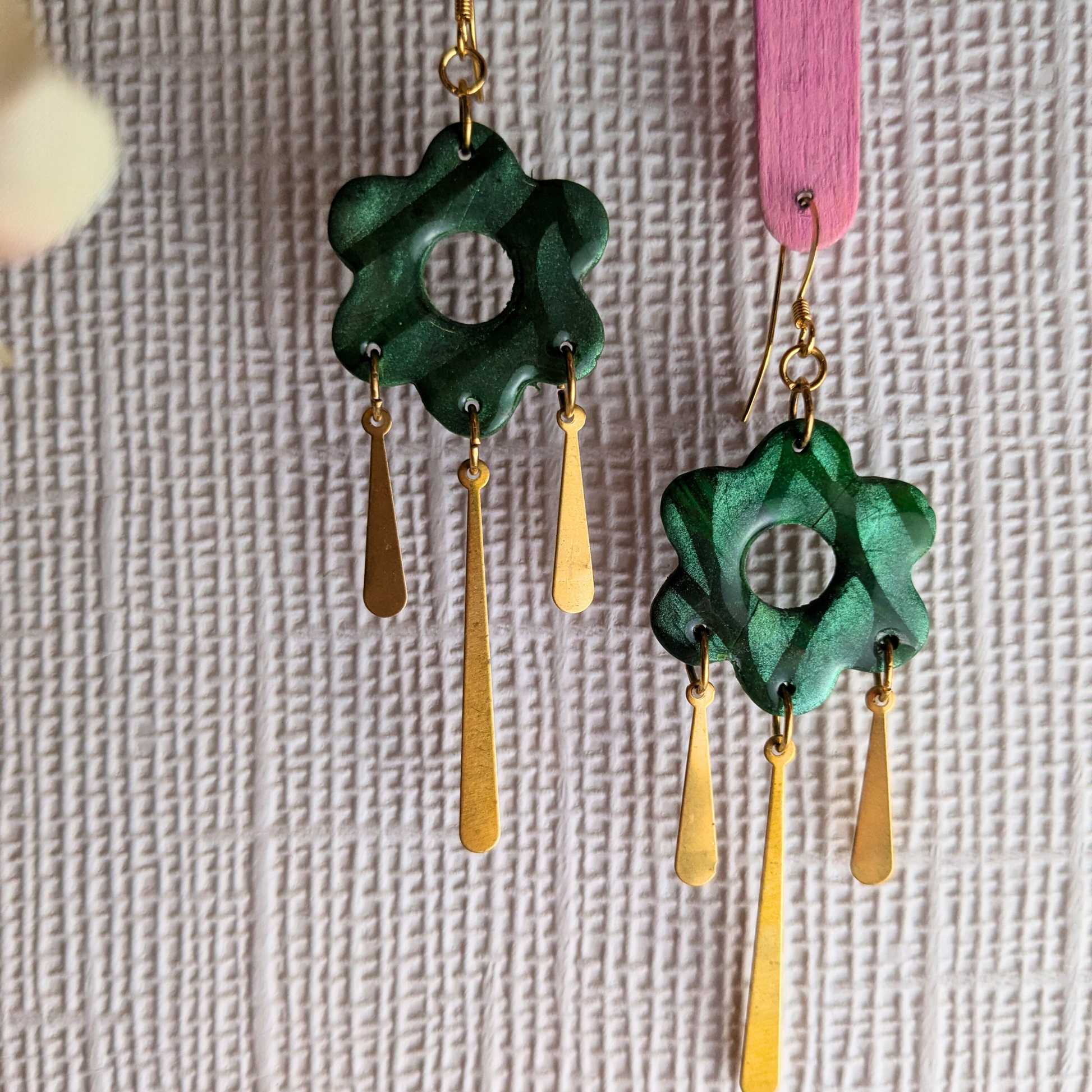 A pair of handmade dangle earrings featuring textured dark green flower-shaped pendants with gold-tone hooks and three dangling gold-tone teardrop charms, shown against a textured light gray background.