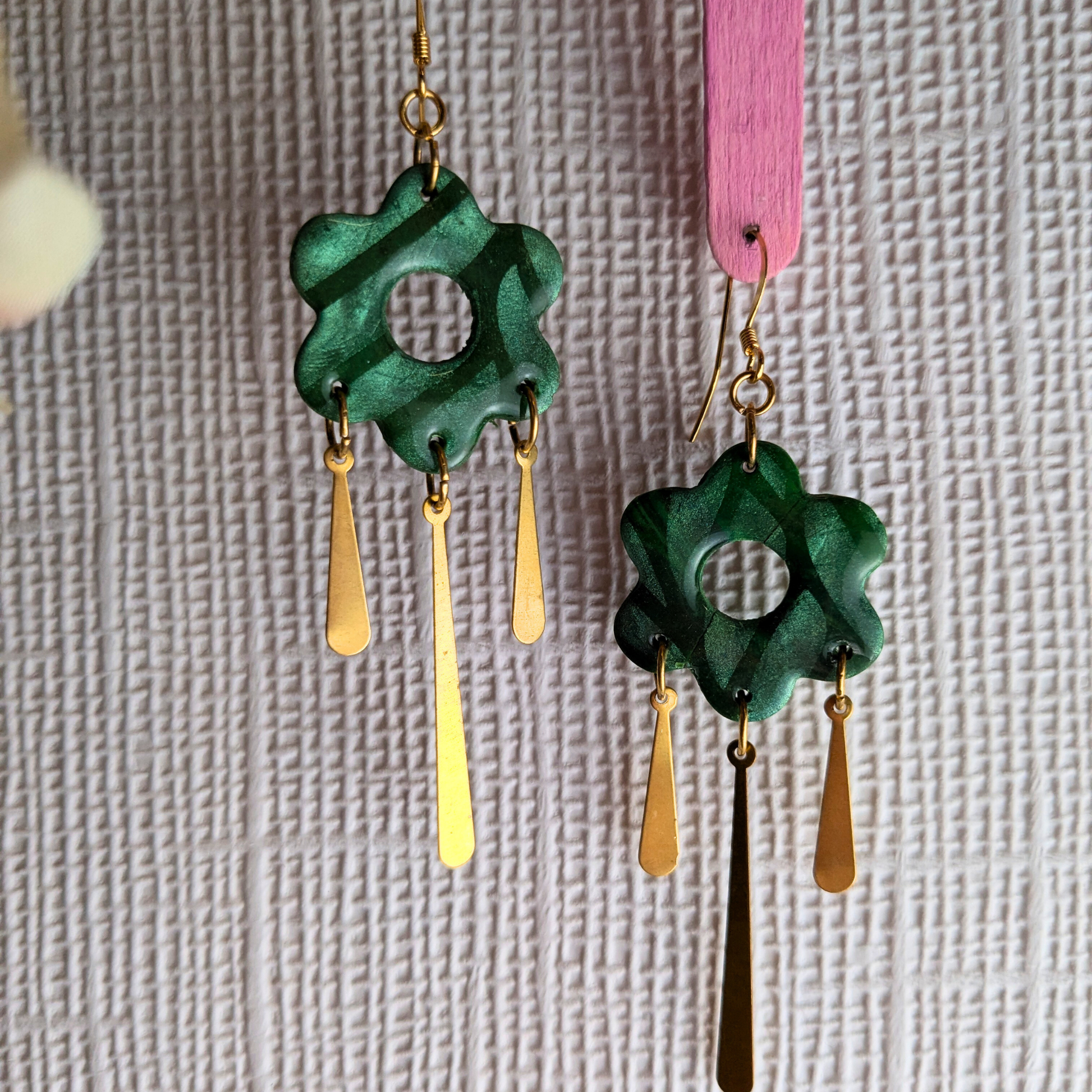 A pair of handmade dangle earrings with textured green flower-shaped charms and gold-tone teardrop pendants, hanging against a textured white background.