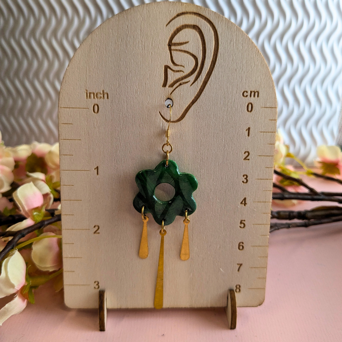 A pair of dangling earrings displayed on a wooden ear-shaped measuring stand. The earrings feature a dark green flower-shaped charm with a circular cutout and two gold-tone dangling tassels.