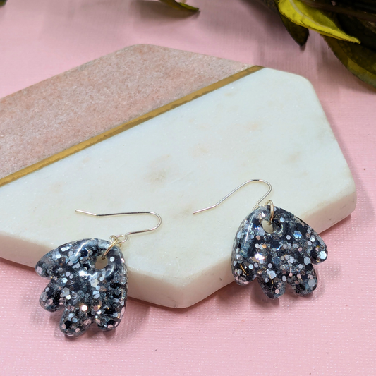 Glittery silver-flake earrings shaped like paws on a hexagonal marble tray. The tone is playful, set against a light pink background.