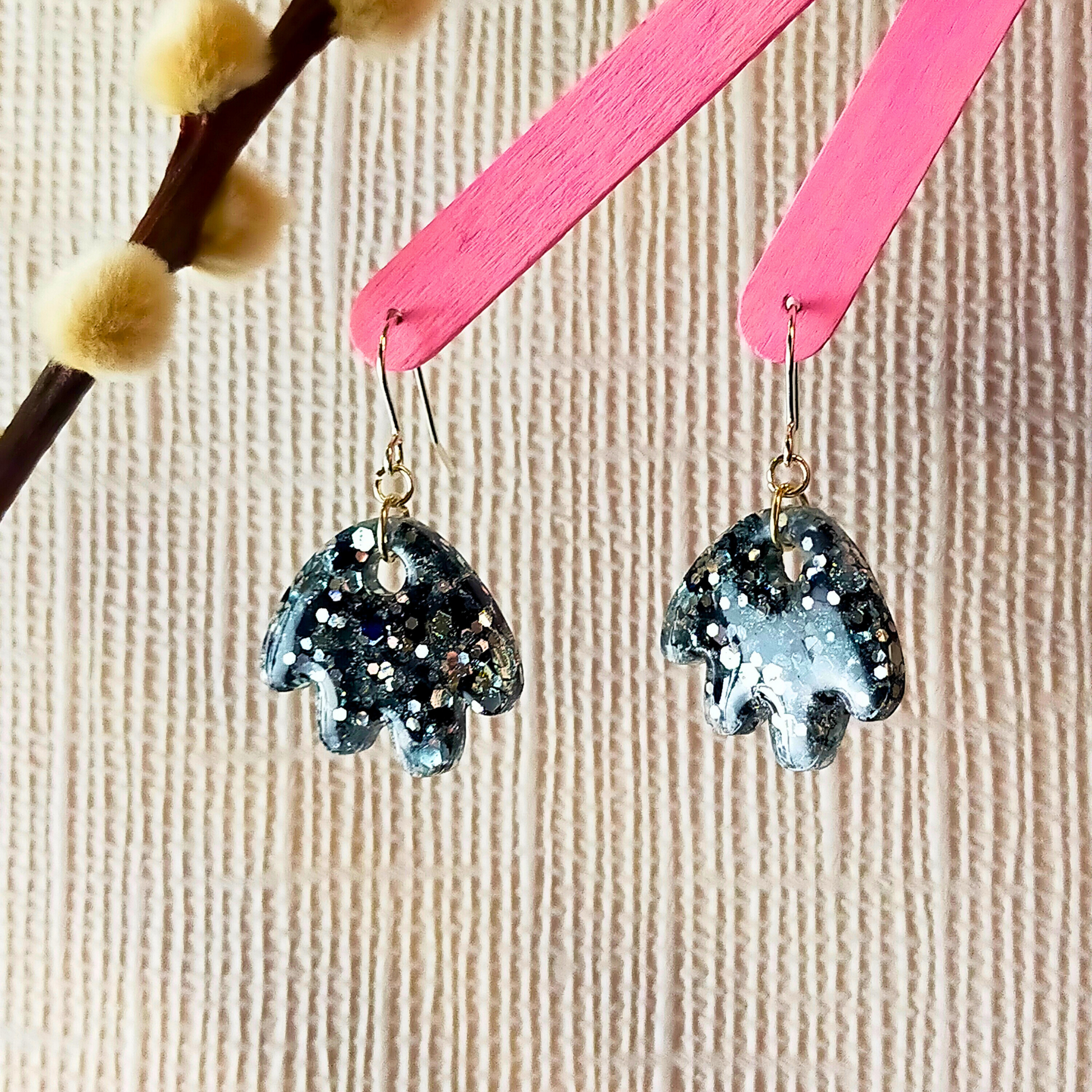 Sparkling black resin earrings with silver glitter hang from pink sticks, set against a textured beige background. A branch with soft buds is visible on the left.