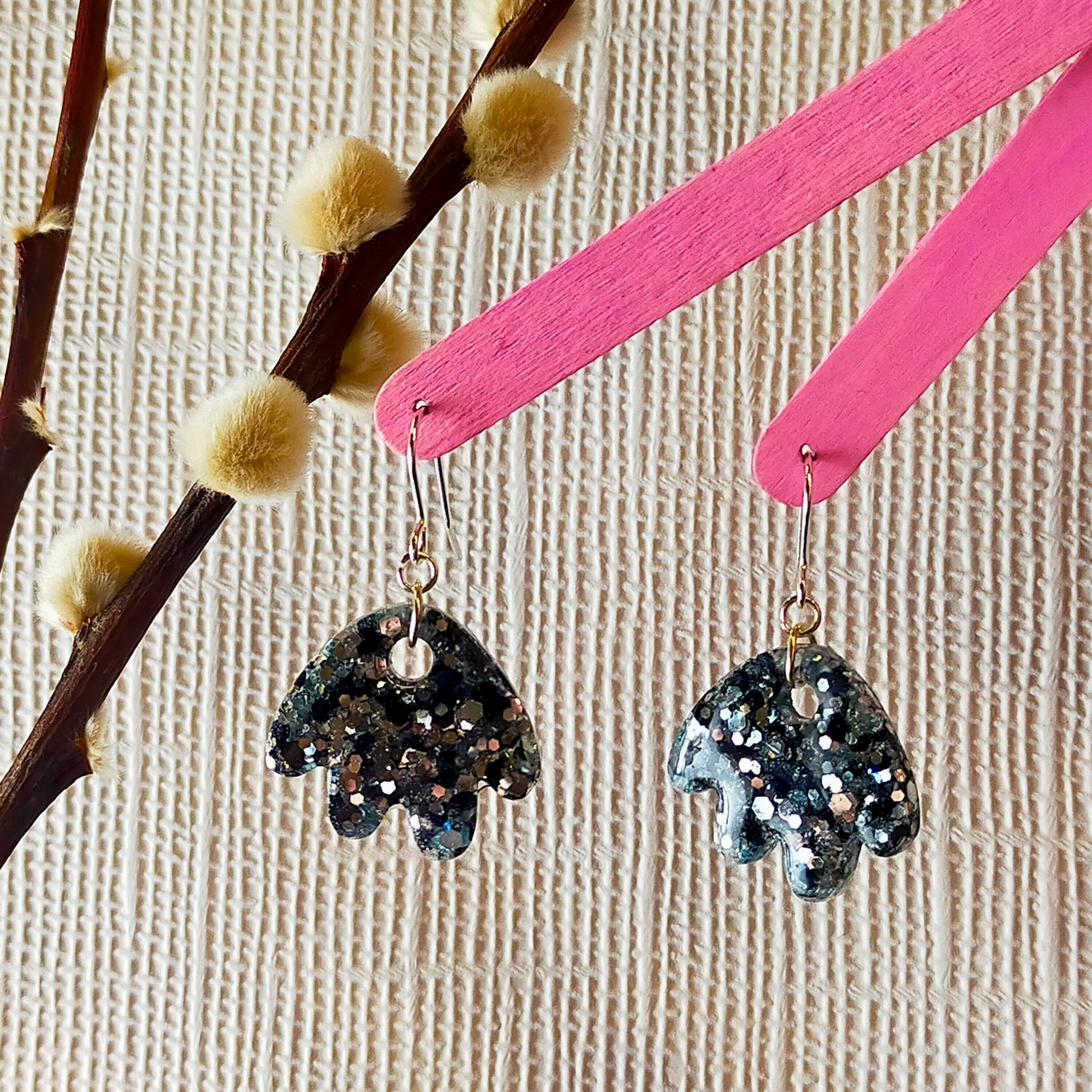 Sparkling black resin earrings with silver glitter hang from pink sticks, set against a textured beige background. A branch with soft buds is visible on the left.