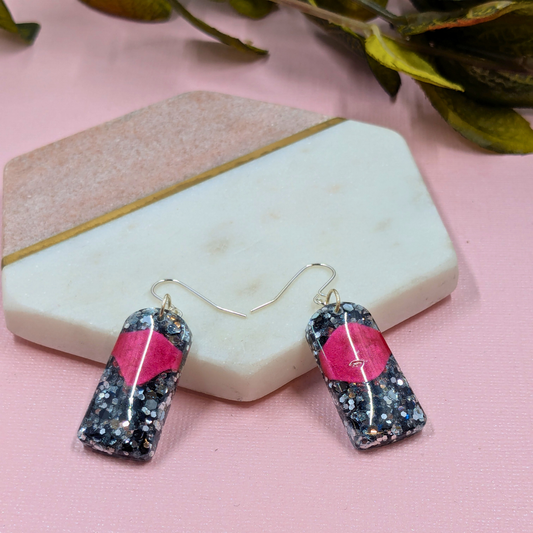 Earrings with black and silver glitter and a red accent rest on a hexagonal stone backdrop. The scene has a stylish, modern tone.
