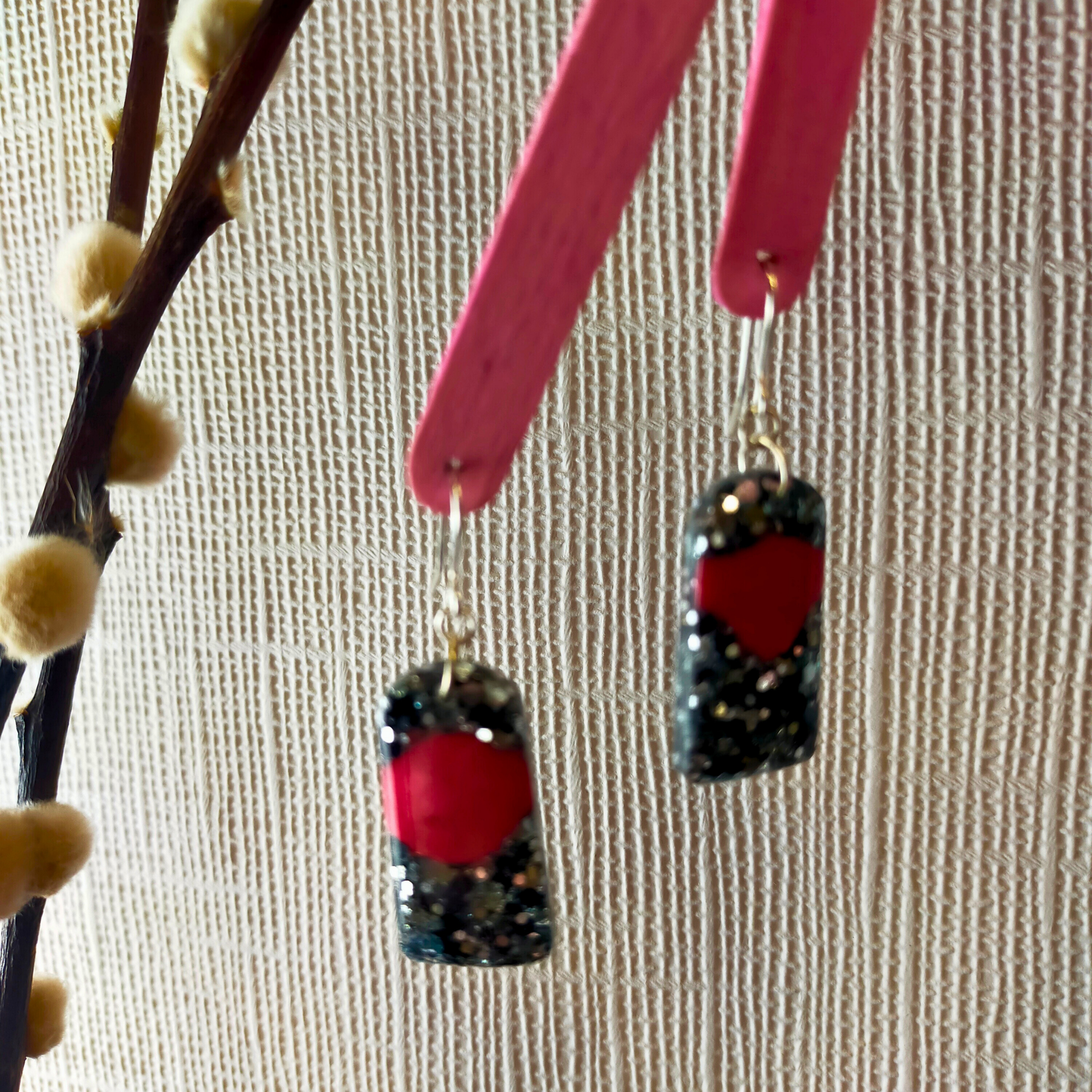 Earrings with black and silver glitter and a red arch dangle from pink holders. They hang near a fabric wall and branches with fluffy buds.