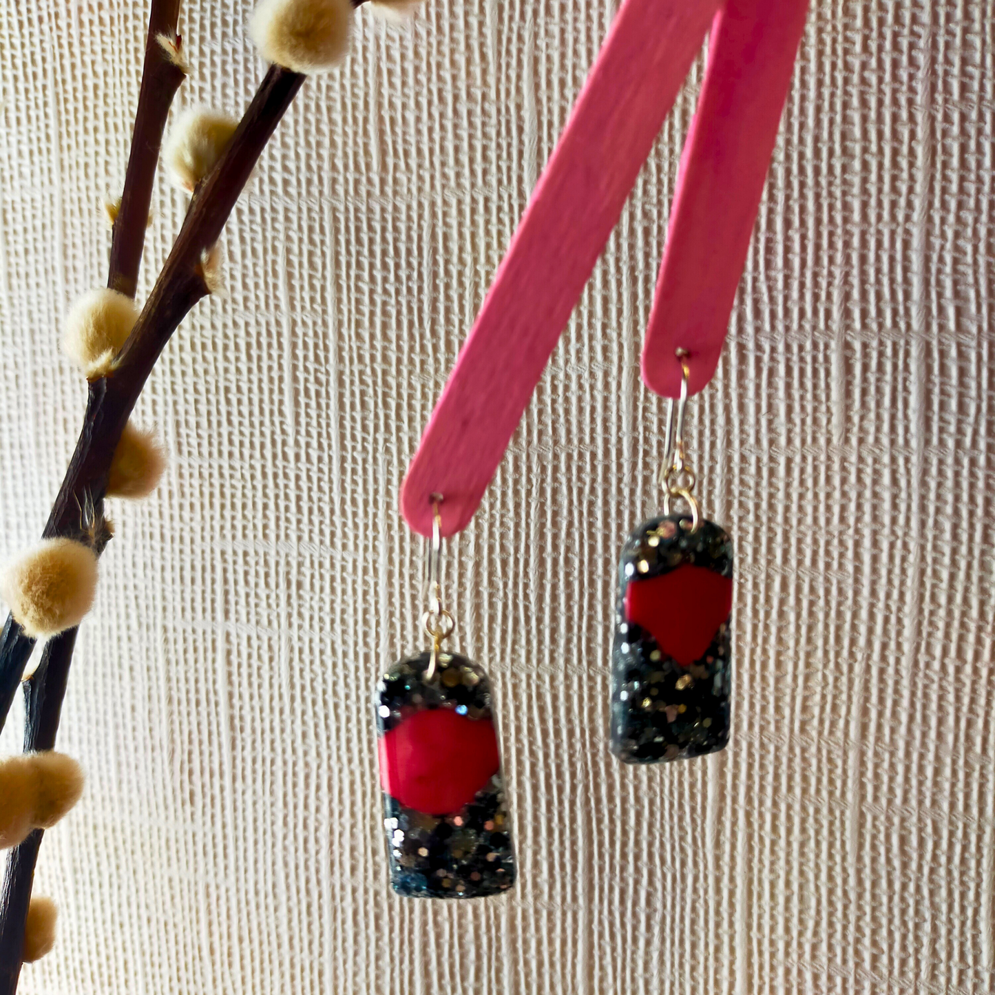 Pink popsicle sticks display glittery black earrings with red ache designs. They hang near a branch with fuzzy buds, against a textured beige background.
