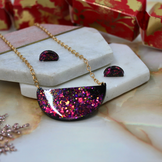 A gold-tone necklace with a glittery semi-circular pendant in shades of magenta, purple, and pink, displayed with matching stud earrings on white stone blocks.