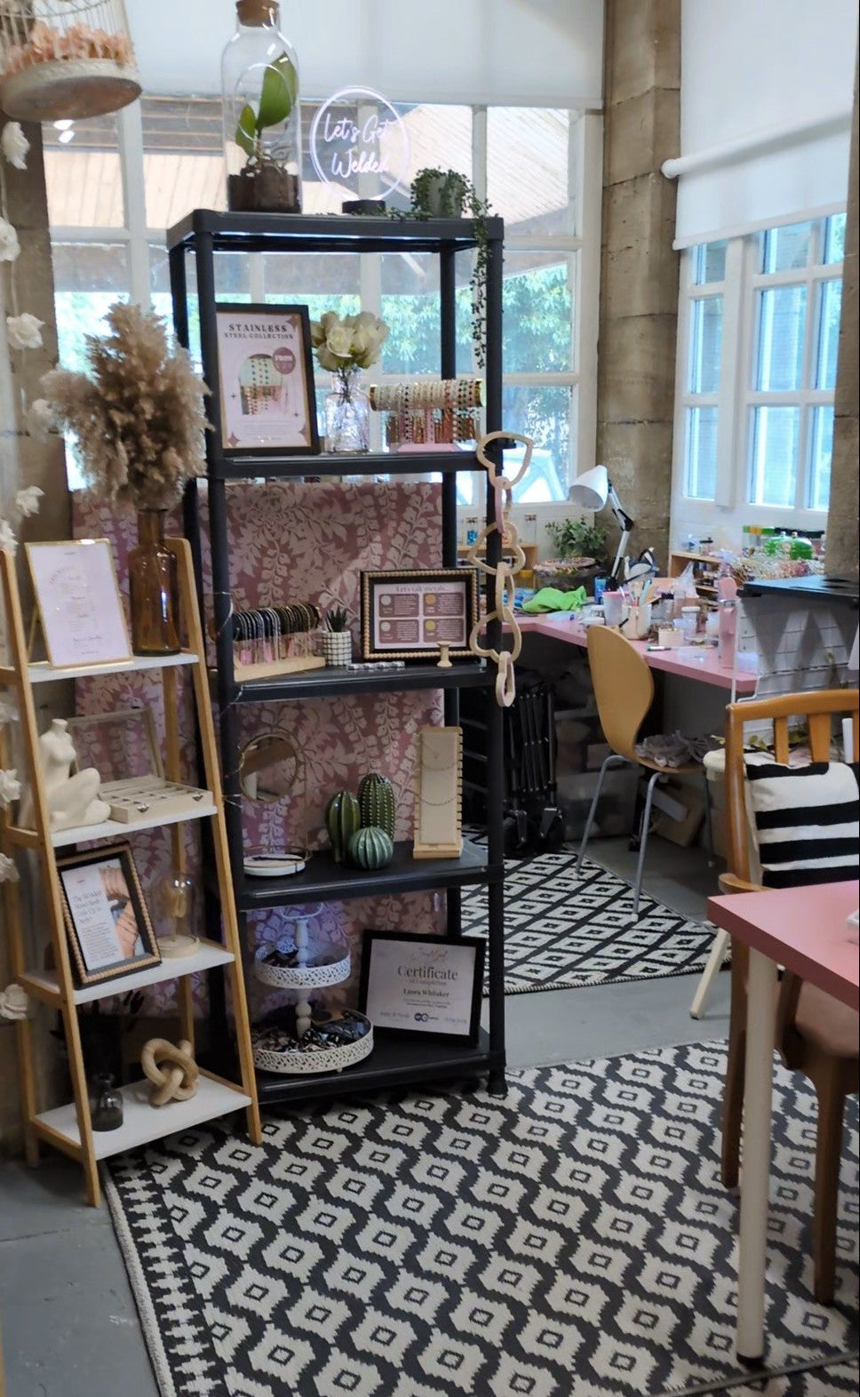 Created By Lau and Welded Bonds Studio - a styled interior scene showing a black metal shelving unit with decorative items, a wooden ladder shelf, patterned rugs, and furniture in a bright room with large windows.