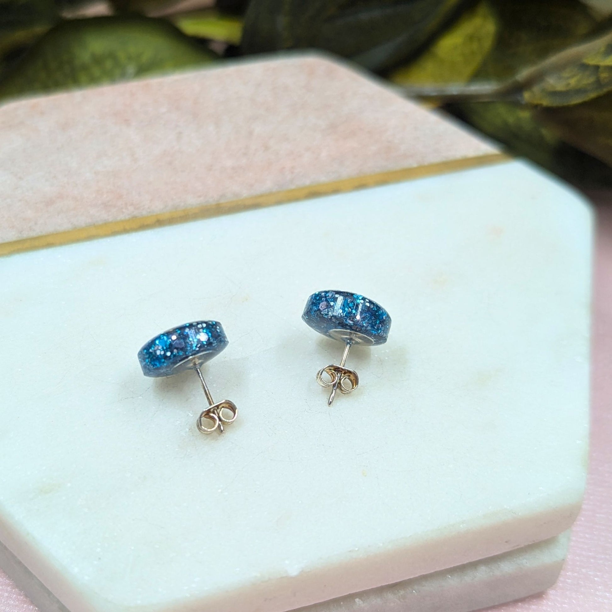 Blue gemstone stud earrings on a white jewelry display box with green leaves in the background.