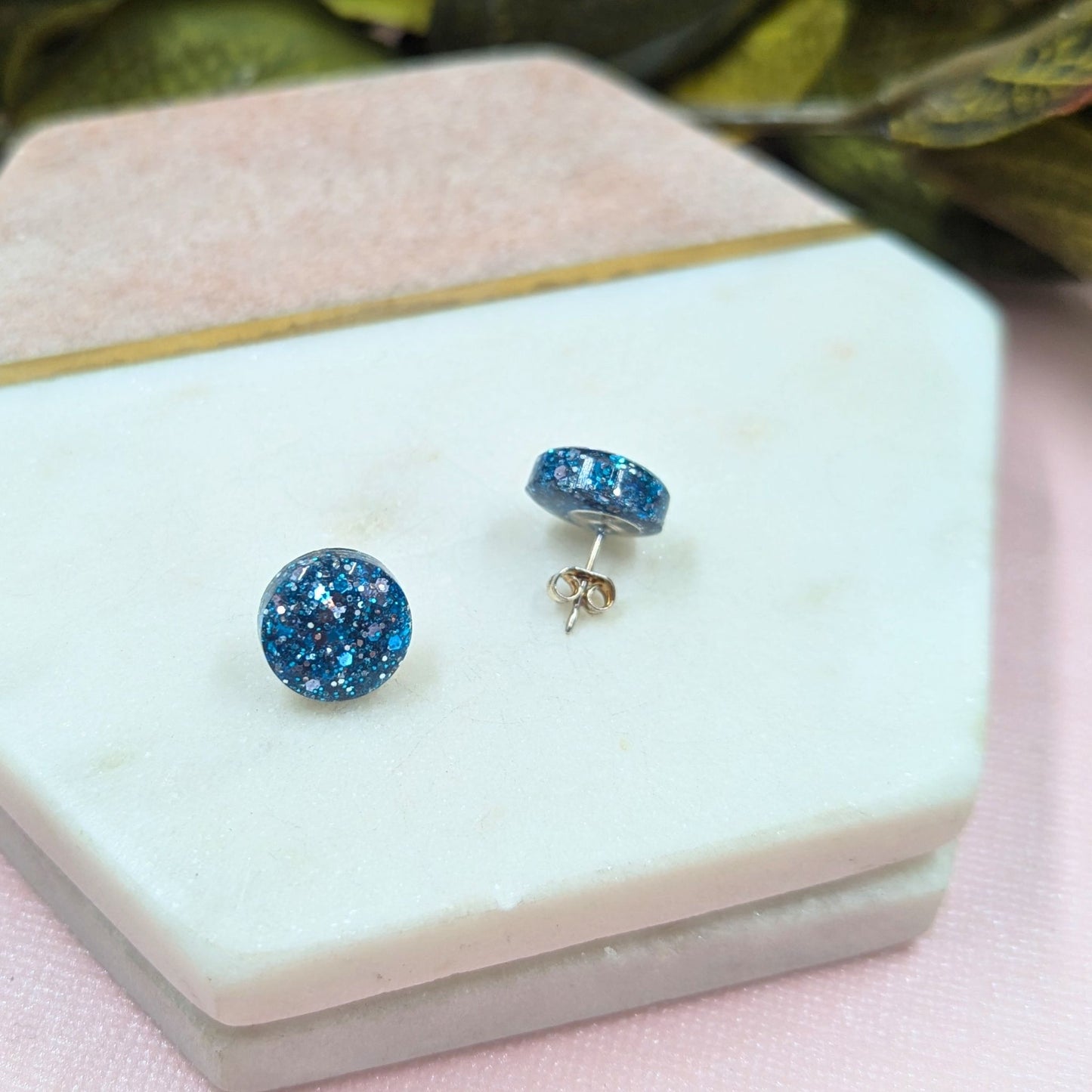 Blue glittery stud earrings on a white jewelry box with a pink surface and green leaves in the background.